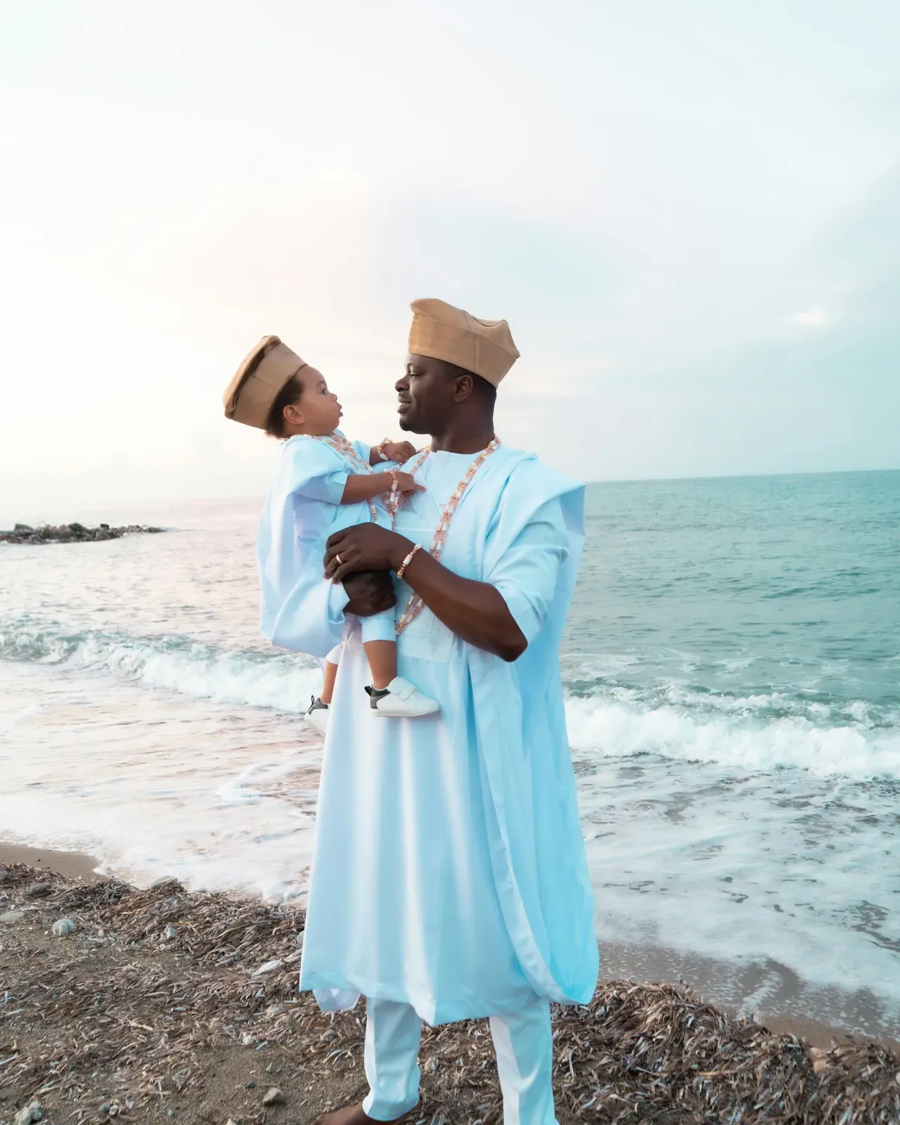 Mann in einem weiten, blassblau-weißen Agbada mit passender Kappe, der seinen kleinen Sohn im Arm hält, der ebenfalls in Weiß mit Kappe gekleidet ist, an einem Strand