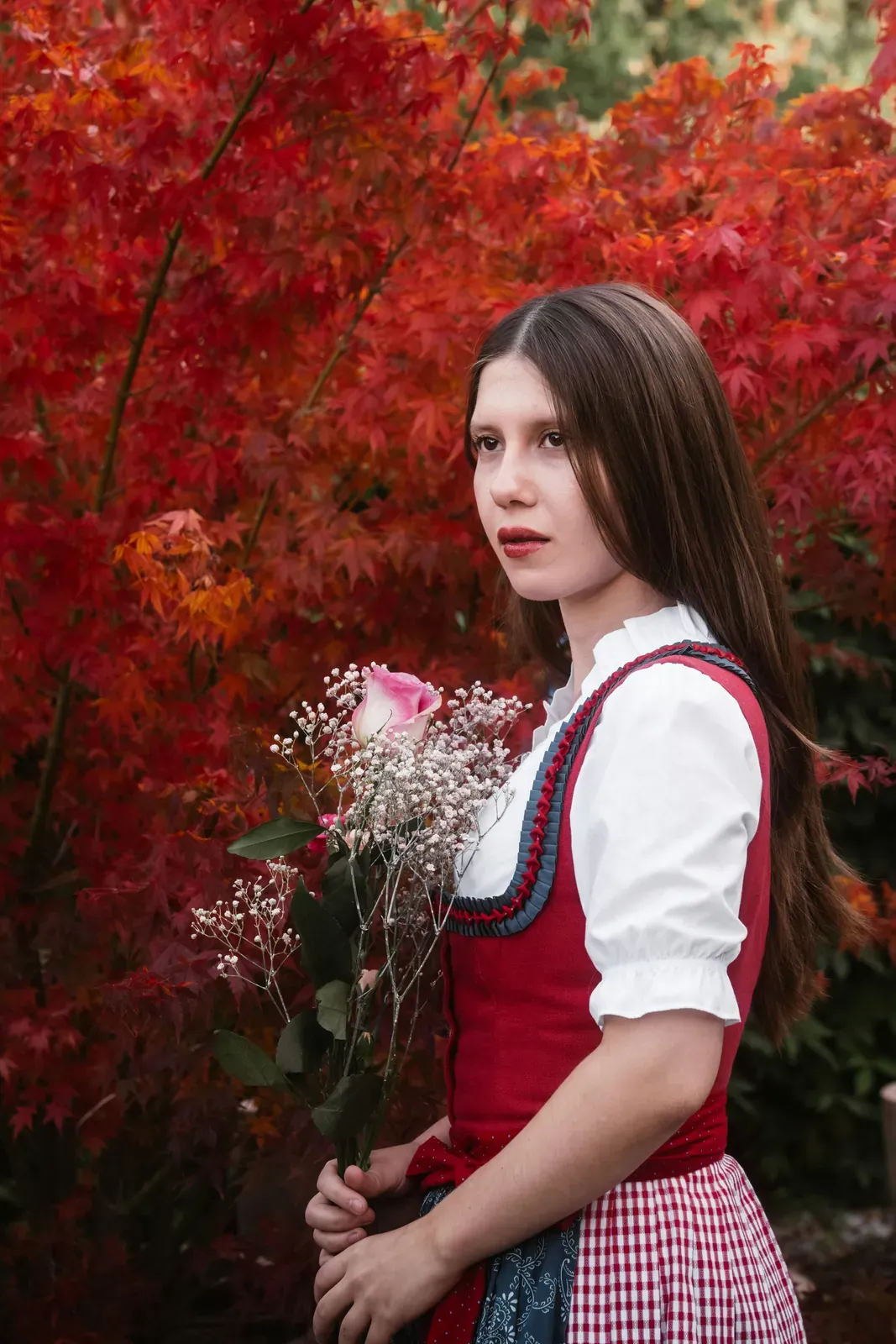 Junge Frau im Dirndl mit rotem Mieder, weißer Puffärmelbluse und rot-weiß karierter Schürze, in der Hand ein Strauß mit rosa Rose und Schleierkraut, dahinter rotes Herbstlaub