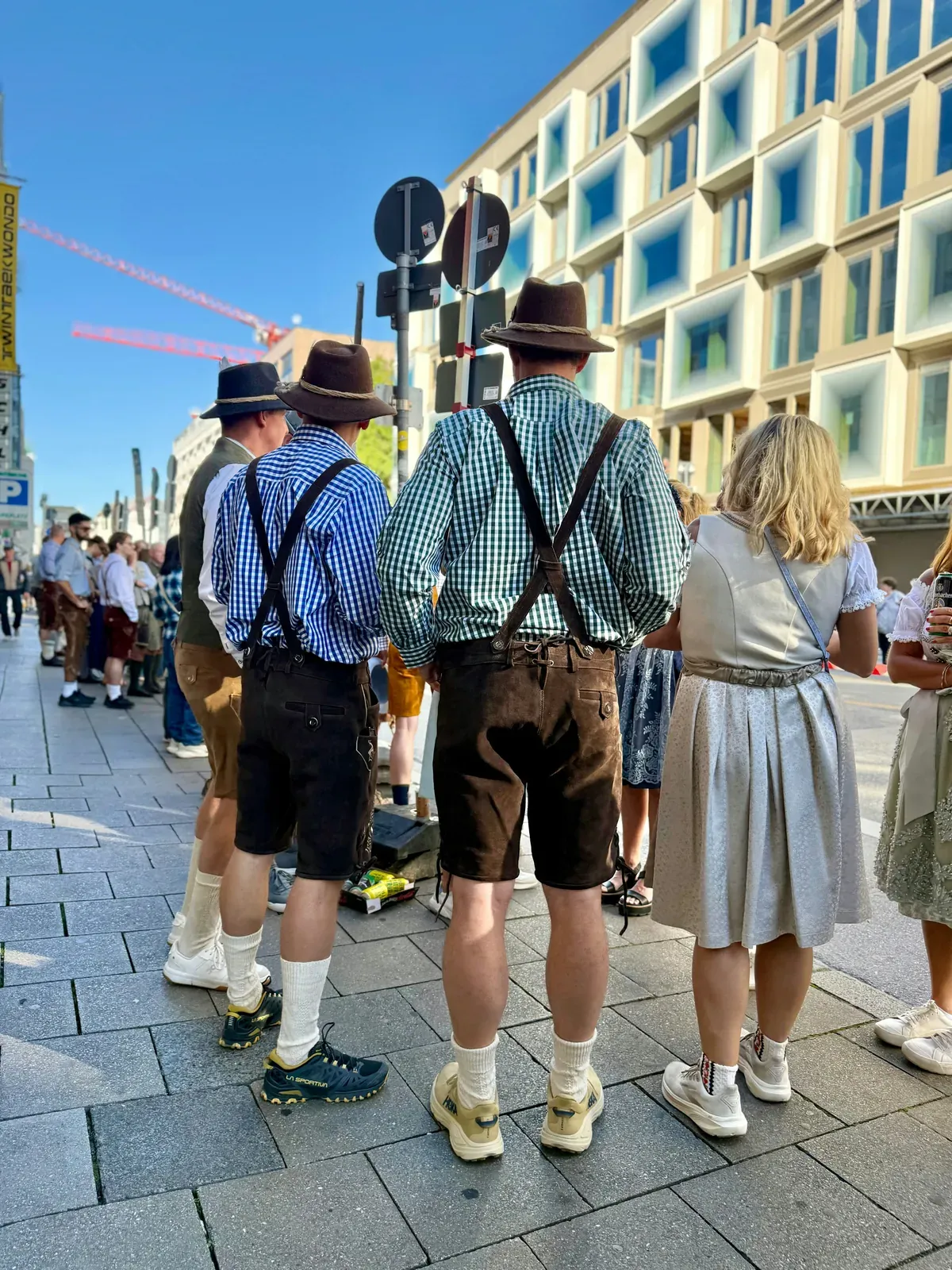 Menschen von hinten auf einem sonnigen Gehweg in München, Männer in Lederhosen mit Hosenträgern, karierten Hemden und Filzhüten, dazu eine Frau im hellen blaugrauen Dirndl