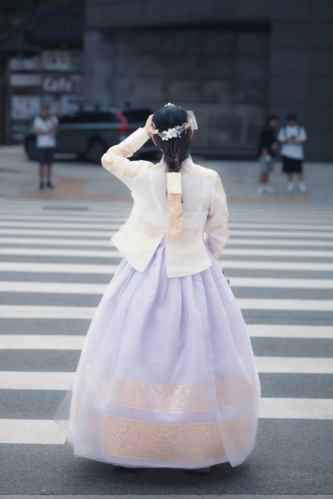 Frau von hinten auf einem Zebrastreifen in Seoul, in einem pastellfarbenen Hanbok mit cremefarbener Jacke, weitem lavendelfarbenem Rock und Blumen im Haar