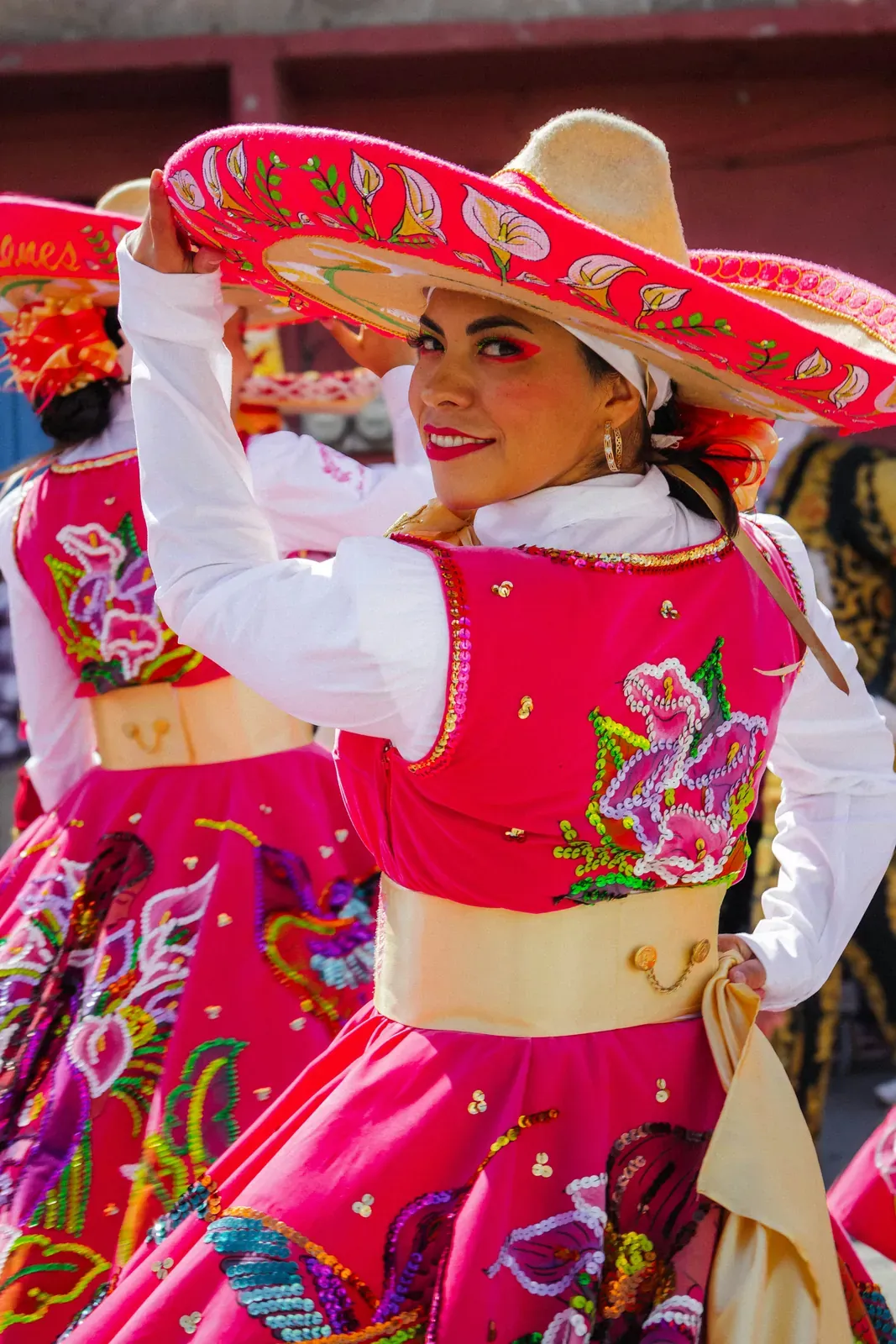 Lächelnde Frau bei einer festlichen Tanzvorführung in einem leuchtend pinkfarbenen Kleid mit großen bunten Blumenstickereien, weißen Ärmeln und gelber Schärpe, die einen breiten rot-weiß bestickten Sombrero hochhält
